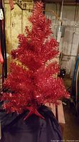 Red tinsel Christmas tree on a base, displayed in an indoor setting.