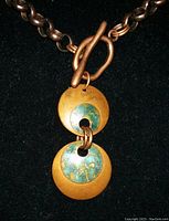 Close-up of a handcrafted necklace with two round copper pendants showing patina and a toggle clasp on a chain, set against black fabric.