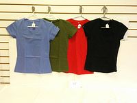 Four T-shirts in mauve, khaki, red, and black hung on a wall rack.