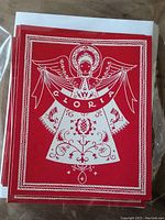 Front view of a red vintage Christmas card designed by Dora De Pedery-Hunt featuring a white folk-Baroque style angel holding a banner with the word 'GLORIA'. The card has a decorative pattern border.