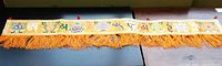 Full view of Tibetan silk brocade wall banner measuring 51 inches long, vibrant yellow with orange fringe tassels along bottom edge and red hanging loops at top edges.