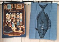 Two cotton tea towels hanging side by side; left one blue with fish print and text, right one colorful detailed illustration of fish and chips shop.
