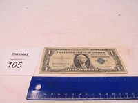 One dollar 1957 A blue seal silver certificate lying flat on a white surface with a ruler for scale