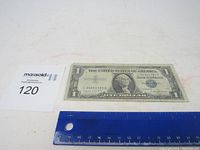 Top down photo of the 1957 A Silver Certificate $1 note with blue seal placed next to a ruler and a lot number card for size reference.