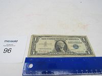 Front side of the 1957B $1 United States Silver Certificate with blue seal next to a ruler for size reference.