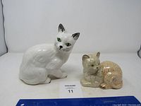 Lot contains two ceramic cat figurines; larger white cat sitting upright, smaller beige resting cat with crackled glaze.