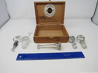 Wooden Ashton cigar box open with five wine bottle stoppers and a metal bottle stopper in front, ruler included for scale.