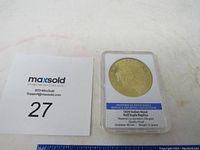 Photo of the replica gold coin in a clear plastic case alongside a MaxSold lot number card showing number 27.