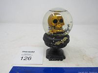 Front and side close-up view of gold skull inside the snow globe on a black pedestal with visible paint chipping and wear.