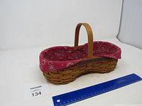 Longaberger peanut-shaped basket with handle and red paisley fabric liner inside, shown at an angle with a blue ruler for size reference.