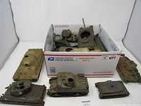 Photo showing a box containing various military tank models along with some tank models placed outside the box. The tanks vary in model, color and size.