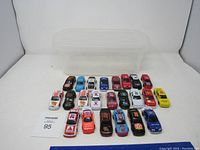 Overhead view of 23 miniature Nascar toy cars arranged in rows in front of a clear plastic storage container.