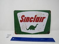 Frontal view of the rectangular metal Sinclair gas sign showing the full logo with green border and dinosaur icon, placed next to a ruler for scale.