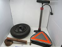 Photo showing used tire with metal hub, manual air pump in silver and black with red base, reflective emergency triangle in carrying case, and rusted L-shaped metal tool.