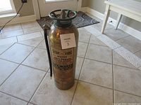 Full view of the vintage brass fire extinguisher showing cylindrical brass body, attached hose, and label with instructions.