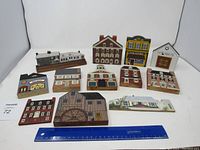 Photo of 12 flat wooden building fronts laid out, featuring mixed styles including shops and houses, in various colors and designs. A ruler is placed in front for size reference.