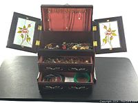 Open wooden jewelry box displaying stained glass doors with floral design, hooks for necklaces, and velvet-lined compartments with many pieces of jewelry inside.