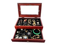 Wooden jewelry box with top glass lid and a drawer filled with various costume jewelry including brooches, bracelets, earrings, and pins.