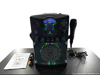 Front view of karaoke machine showing microphone, cables, instruction manual, and colorful lit speaker grille.