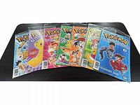 Seven Pokémon comic books displayed fanned out on a black surface, showing colorful covers featuring Pikachu and main characters from the series. Each comic is in a clear protective sleeve.