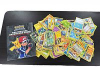 Photo of official Pokémon Adventure Guide book with cover featuring Ash and Pikachu alongside a large pile of Pokémon trading cards spread out.
