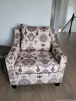Front-left angled view of the armchair showing the fabric upholstery with decorative patterns in beige, purple, and dark brown tones, cushioned seat and back, and dark wooden legs.