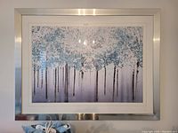 Framed painting of a forest with tall trees and blue-tinted leaves in silver frame, hung on wall.