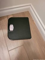 Photo showing a black mechanical bathroom scale with analog dial on a wood floor corner.
