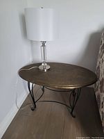 Oval metal coffee table with textured bronze-gold top and black wrought iron legs shown with lamp on top.
