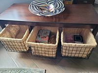 Photo of the dark wood rectangular coffee table with three black wire frame baskets with beige fabric liners underneath. A swirl pattern glass decorative bowl sits on top but is excluded from the lot.