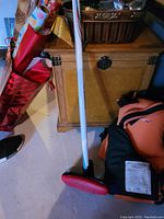 Photo showing a white curling stick with black grip, black head with red pad, positioned next to a black and orange curling glove and an orange and black carry bag