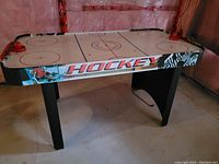 Full view of air hockey table with hockey-themed graphics and black legs, showing playing surface and score keepers.