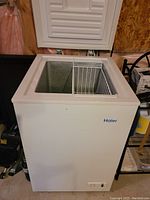Haier small chest freezer with hinged lid open showing interior compartment and metal storage basket.