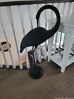 Black metal flamingo sculpture with neck curved downward on wooden floor near white railing.