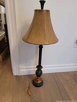 Table lamp with wooden textured lampshade and decorative dark base with carved details and electrical cord coiled at base.