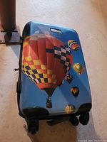 Exterior of blue hard-shell suitcase featuring colorful hot air balloon graphic, four wheels and bottom handle showing damage