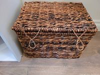 Large rectangular woven wicker storage basket with lid and loop handles sitting on a wooden floor.