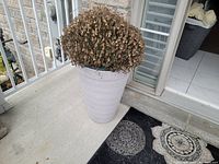 Large gray planter with dried old mum plant placed outside on a concrete porch near entrance door with outdoor rug partially visible on floor.
