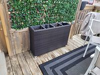 Outdoor dark brown rattan planter with three rectangular compartments, each filled with soil. Positioned on a wood deck next to wooden wall and chair.