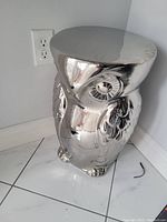 Silver owl-shaped accent table with reflective finish placed on a white tiled floor beside a wall outlet.