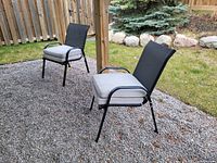 Two black outdoor chairs with metal frames and mesh backs and gray cushions installed, pictured on gravel with grass and wooden fence background.