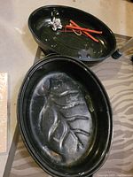 Black oval roasting pan with ribbed interior and matching lid with handle, displayed open on a surface with few small items inside lid.