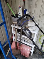 Garden tools and cleaning supplies including shovel, pink snow shovel, sprayer bottle, storage bucket, broom, and yellow garden hose coiled.