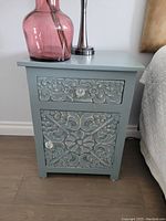 Light blue wooden side table with carved drawer and door, floral design, lamp and vase on top