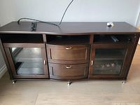 Front view of a dark brown wooden TV entertainment unit with two curved drawers in the center and two glass doors on the sides showing interior shelving. There are open compartments on top of the drawers and doors.