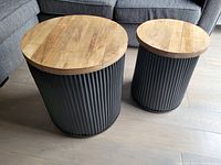 Photo shows two decorative end tables with round natural wood tops and black vertically ribbed cylindrical bases, placed on a wooden floor in front of a grey couch.