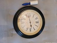 First clock with black frame, white aged dial, Roman numerals and black hands, lint roller for scale.