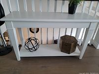 White wooden console table with a lower shelf, placed against a white railing indoors, displaying two decorative items on the bottom shelf.