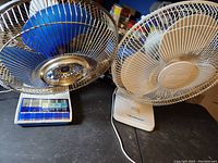 Photo showing both vintage oscillating fans side by side on a surface. One fan with blue blades and blue and white base, the other with beige blades and beige base.