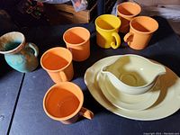 Multiple Fiesta ware and similar ceramic dishware pieces on black surface including orange and yellow mugs, creamy yellow plates and gravy boat, turquoise pitcher, and divided serving dish.